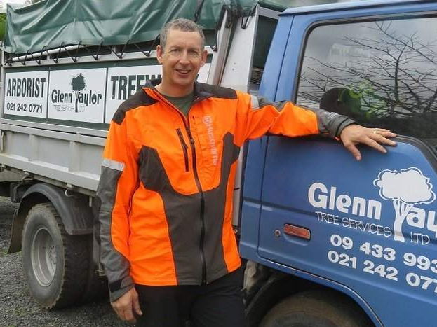Arborist in an orange and grey chainsaw jacket leaning on the door of the blue Glenn Taylor Tree Services truck, with the chipper hitched behind